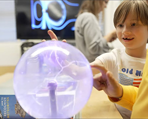 A young child touches a plasma globe, causing bright electric filaments. An adult with a yellow sleeve also engages with the globe, creating a shared moment of curiosity.