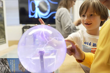 A young child touches a plasma globe, causing bright electric filaments. An adult with a yellow sleeve also engages with the globe, creating a shared moment of curiosity.