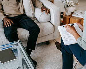 Legs of man seated on beige couch across from female with clipboard in a chair