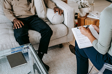 Legs of man seated on beige couch across from female with clipboard in a chair