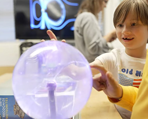 A young child touches a plasma globe, causing bright electric filaments. An adult with a yellow sleeve also engages with the globe, creating a shared moment of curiosity.