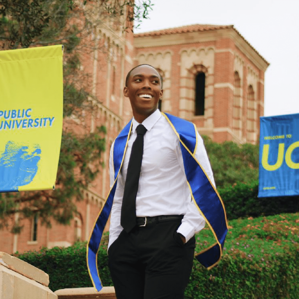 A smiling graduate in a white shirt, black tie, and blue-and-gold sash stands outdoors near a brick university building with Public University and Welcome to UC banners in the background.