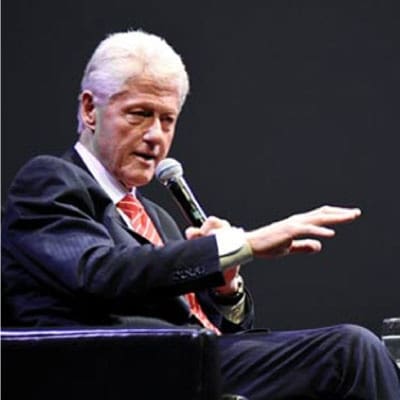 An older man in a suit and red striped tie speaks into a microphone while seated, gesturing with his hand against a dark background.