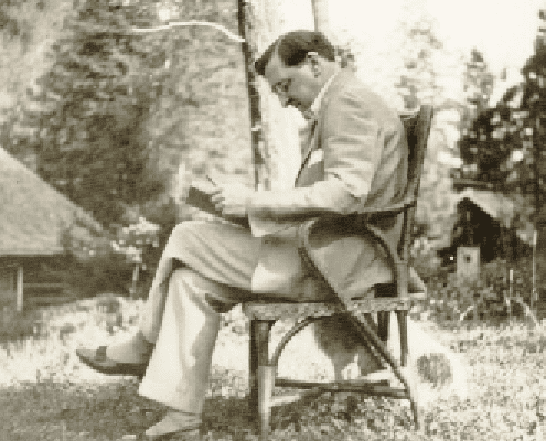 A man in a light-colored suit sits on a wooden chair outdoors, reading a book. Trees and a building are visible in the blurred background, suggesting a peaceful, natural setting.