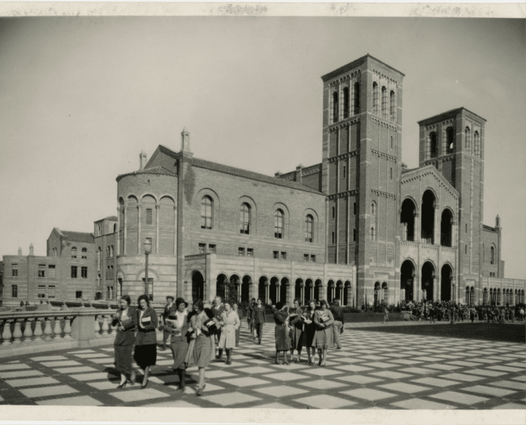 A black-and-white photo of a large brick building with arches and towers, resembling a university hall, with groups of people walking and gathering on a checkered plaza in front.