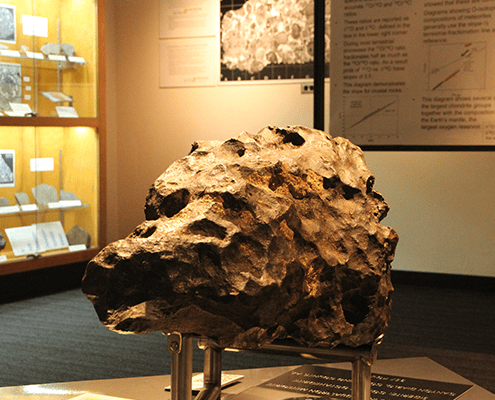 A large, dark, pitted meteorite is displayed on a stand in a museum exhibit, with informational displays and rock samples visible in the background.
