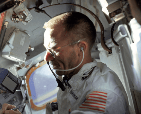 An astronaut in a white suit with an American flag patch, wearing a headset, works inside a spacecraft filled with equipment and controls, with sunlight illuminating his face.