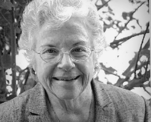 An older woman with curly white hair, glasses, and a warm smile is wearing a blazer and sitting outdoors in front of leafy branches. The image is in black and white.