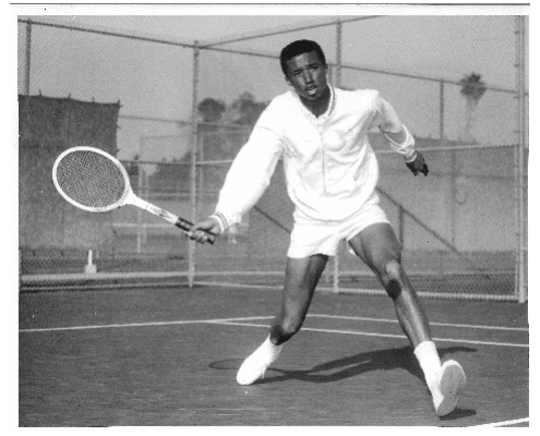 A Black male tennis player in a white outfit lunges forward to hit a tennis ball with his racket on an outdoor court, with a fence and trees visible in the background.