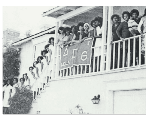 A group of women pose on a staircase and balcony outside a house, smiling at the camera. They hold a banner displaying Greek letters ΔΣΘ (Delta Sigma Theta). The image is in black and white.
