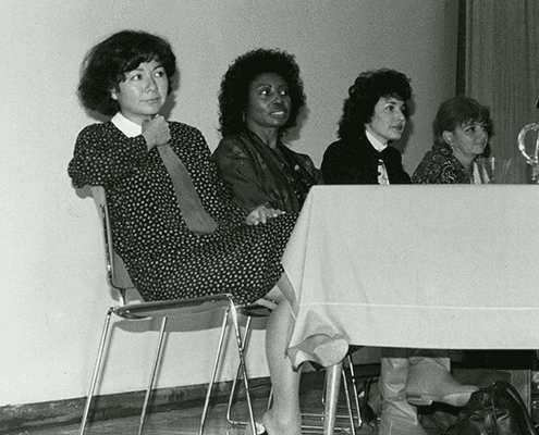 Four women sit at a table during what appears to be a panel discussion or meeting. Three look forward attentively, while one looks to the side. The setting is indoors and the image is in black and white.