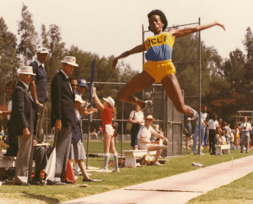 An athlete wearing a UCLA uniform is mid-air during a long jump at an outdoor track event, with spectators and officials watching nearby. Trees and sports equipment are visible in the background.