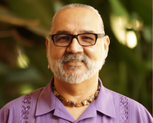 A smiling man with gray hair tied back, a gray beard, and glasses wears a purple shirt and a beaded necklace. Lush green foliage is blurred in the background.