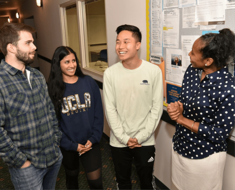Four people stand together in a hallway, smiling and talking. Two are women, one wearing a UCLA sweater, and two are men. They are standing near a bulletin board with papers and notices pinned up.