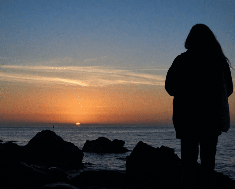 A silhouette of a person standing on rocks by the sea, watching the sun set on the horizon under a sky with soft clouds.