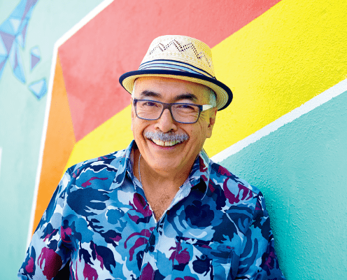 An older man with glasses and a patterned shirt smiles while standing in front of a colorful wall with diagonal stripes of red, yellow, and green. He is wearing a straw hat.