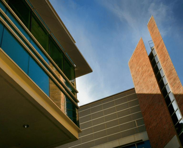 Modern buildings with glass, metal, and brick facades are shown against a clear blue sky. The perspective looks upward, highlighting sharp angles and architectural details.