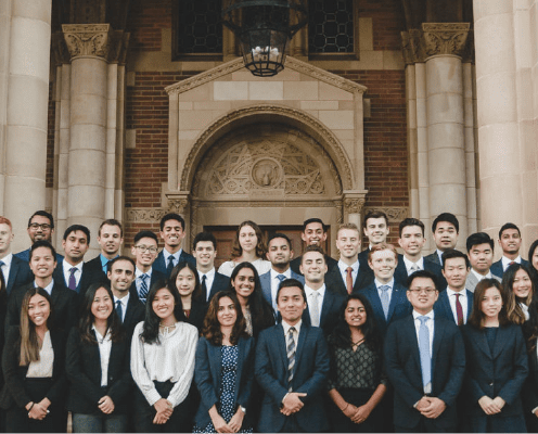 A large group of professionally dressed people, mostly young adults, stand smiling in rows outside a building with ornate stone columns and an arched doorway.