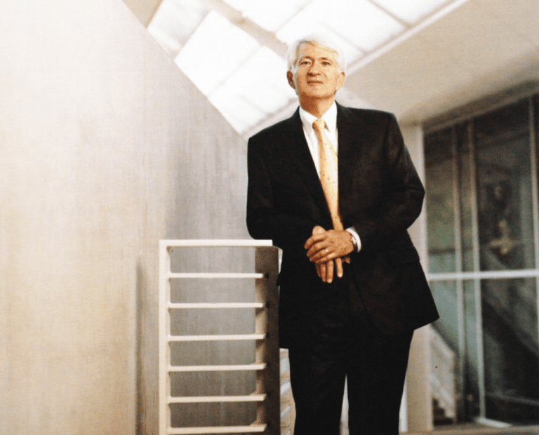 An older man in a dark suit and yellow tie stands indoors by a modern railing, with light streaming in from a skylight above and architectural lines in the background.