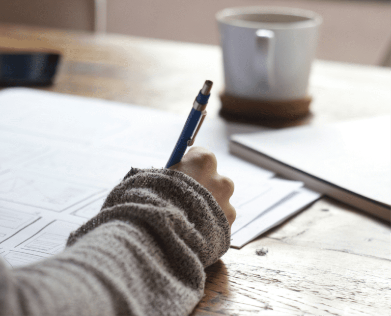 A person wearing a gray sweater writes with a pen on paper at a wooden table. Nearby are a white mug, a closed notebook, and a smartphone.