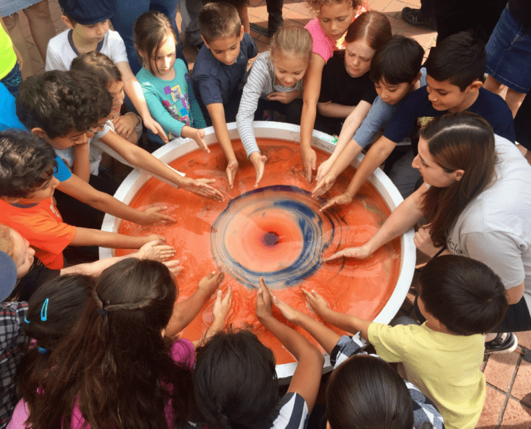 A group of children and an adult stand around a circular table with swirling red and blue liquid, each reaching in to touch it, creating a mesmerizing vortex pattern.