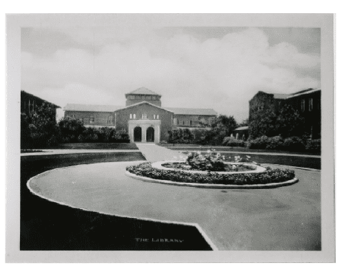 Black-and-white photo of a large brick library building with an arched entry, surrounded by gardens. A circular fountain or flower bed is in the foreground, with a pathway leading to the entrance.