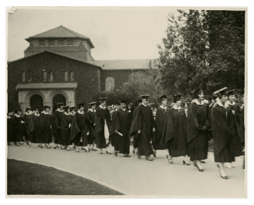 A group of graduates in caps and gowns walk in a procession outside a large brick building, surrounded by trees, on a cloudy day.