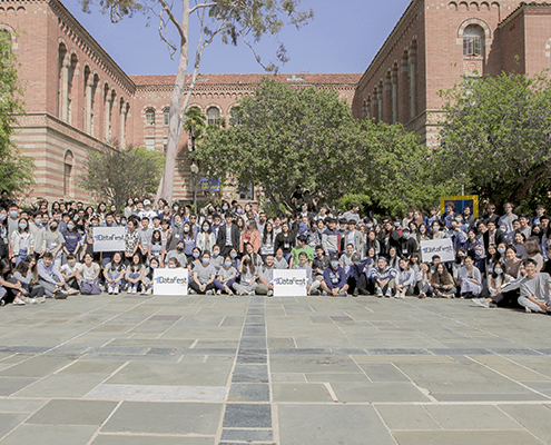 A large group of people poses for a photo outside a brick building, with trees on both sides. Some hold signs reading DataFest. The group is spread out across a stone-paved courtyard under a clear sky.