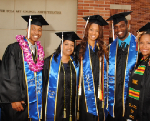 Five graduates in caps and gowns stand together smiling. They are wearing blue stoles, leis, and cords, with a building and UCLA sign in the background.