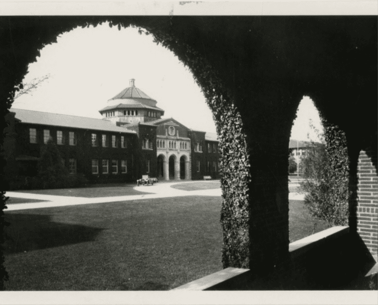 Black and white photo of a large brick building with an arched entrance and a central tower, viewed through a foreground archway. A vintage car is parked in front, and open lawns surround the building.
