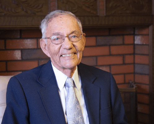 An elderly man with gray hair and glasses, wearing a dark blue suit, light blue tie, and white shirt, sits smiling in front of a brick fireplace.