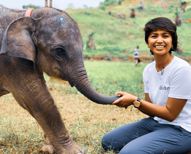 A smiling person kneels on grass, holding the trunk of a baby elephant in an outdoor setting with green hills in the background.