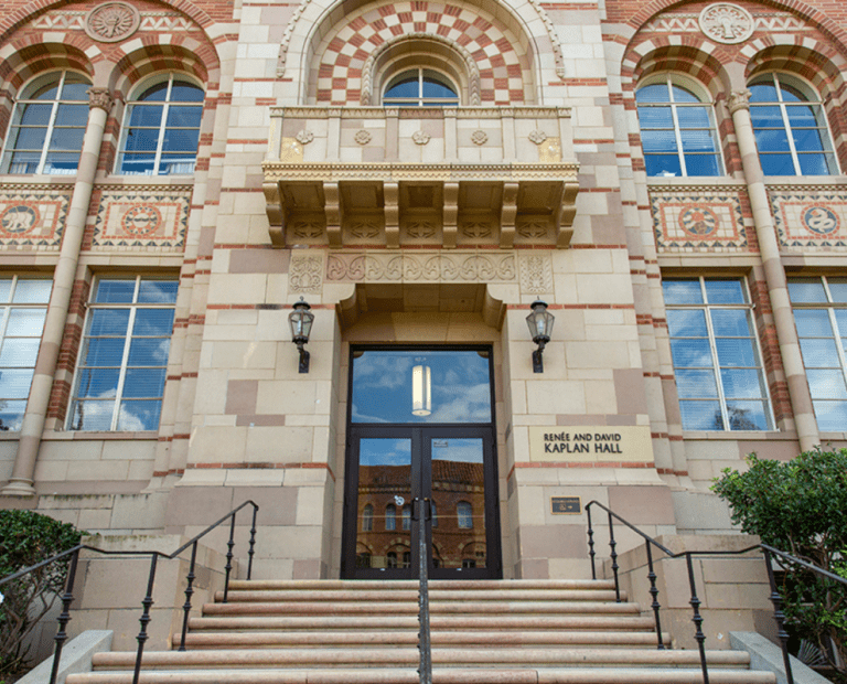 The entrance to a historic building with arched windows, detailed stonework, a balcony, and a sign reading Renée and David Kaplan Hall above the door. Stairs and railings lead up to the door, flanked by greenery.