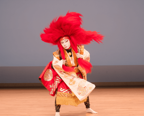 A performer in traditional Kabuki costume with an elaborate red wig and painted face poses dramatically on a wooden stage against a plain background.
