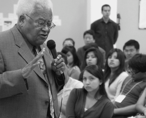 An elderly man in a suit speaks into a microphone in front of an attentive audience of young adults at an indoor event. The atmosphere appears serious and focused.