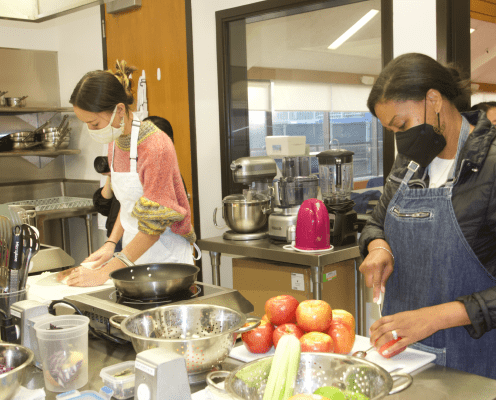Two women wearing aprons and face masks chop vegetables and apples in a kitchen. There are bowls, utensils, and kitchen appliances on the counters around them.