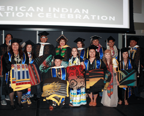 A group of graduates in regalia and traditional Native American clothing stand on stage, holding colorful blankets, smiling, with a screen behind reading American Indian Graduation Celebration.