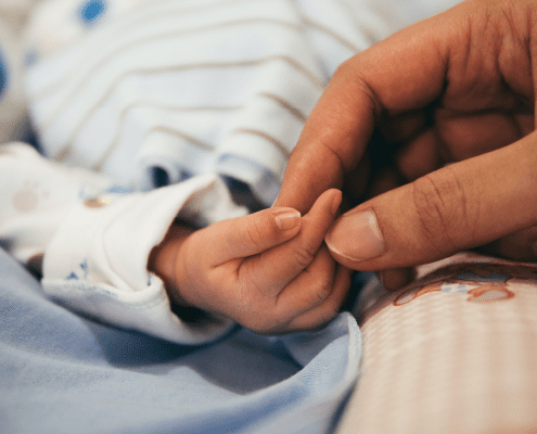 A close-up of an adult gently holding a babys tiny hand, with soft blue and pink fabrics in the background.