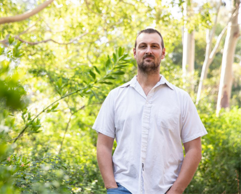 A man with short hair and a beard stands outdoors in a lush, green garden, wearing a short-sleeved white shirt and jeans. Sunlight filters through the trees in the background.