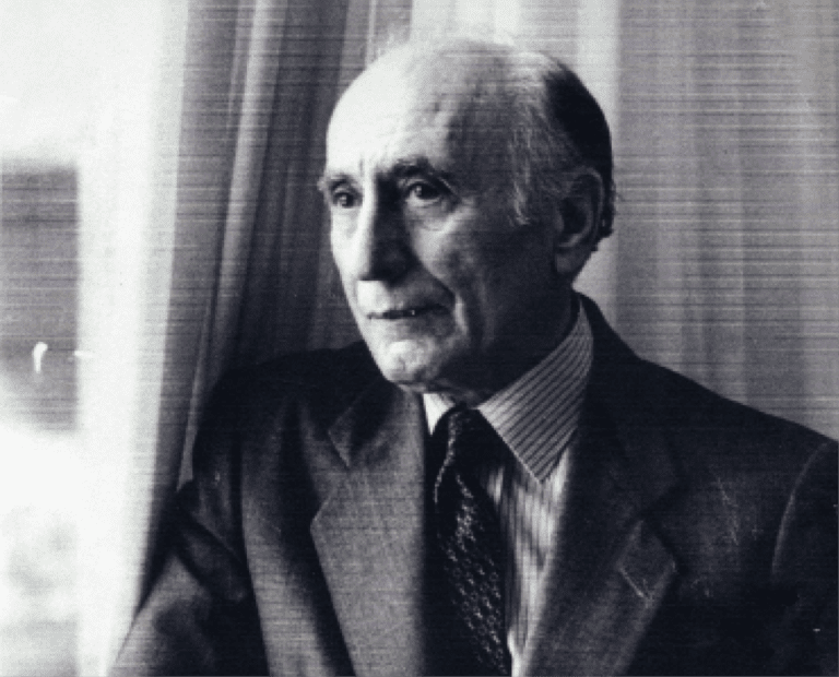 An older man in a suit and tie stands near a window with curtains, looking thoughtful. The image is in black and white.