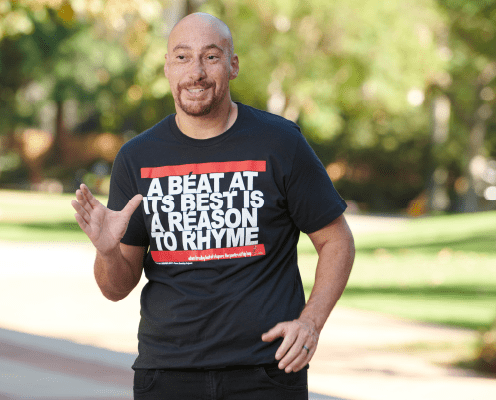 A smiling man with light facial hair stands outdoors, wearing a black t-shirt that says, A beat at its best is a reason to rhyme. Trees and greenery are visible in the blurred background.