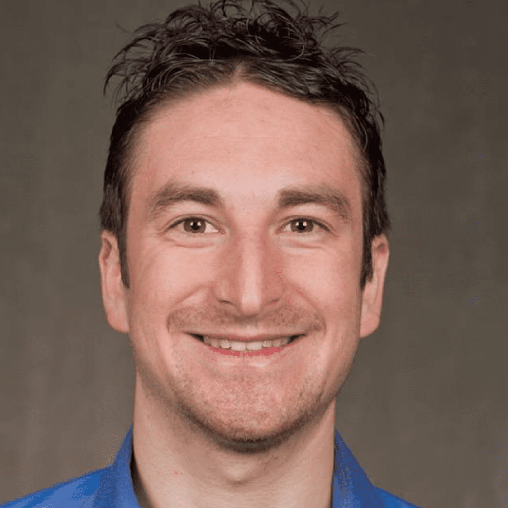 A man with short, dark, slightly curly hair is smiling at the camera. He is wearing a blue collared shirt and is posed against a plain, neutral background.