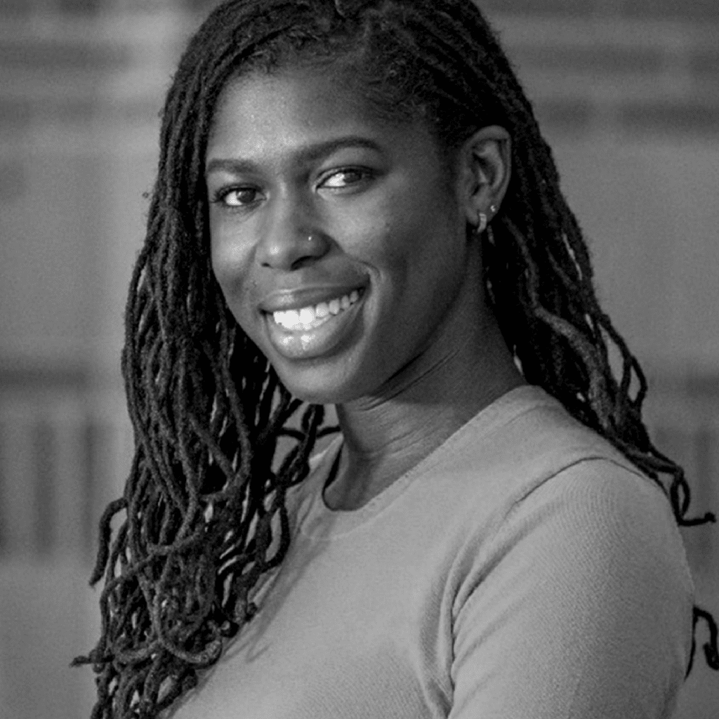 A woman with long, braided hair smiles while looking at the camera. She is wearing a long-sleeved shirt and stands in front of a blurred background. The image is in black and white.