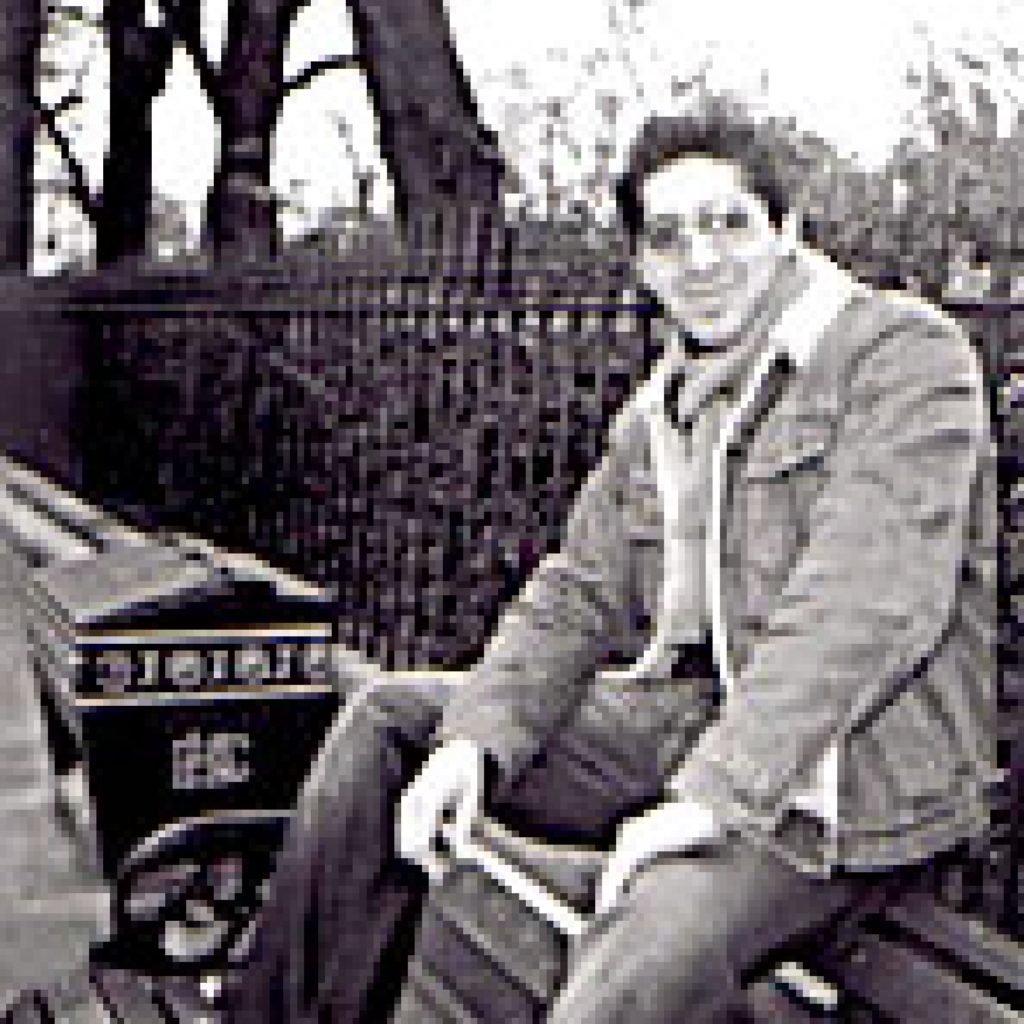 A person wearing glasses and a denim jacket sits on a bench outdoors next to a decorative box, with trees and a metal fence in the background. The image is in black and white.