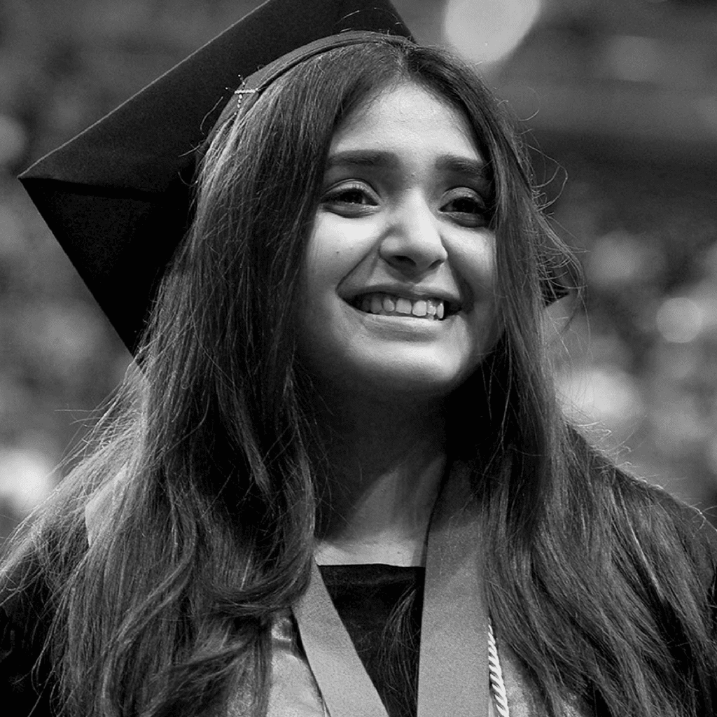 A young woman in a graduation cap and gown smiles brightly, with long hair draped over her shoulders. The background is blurred, suggesting a celebratory event or ceremony.