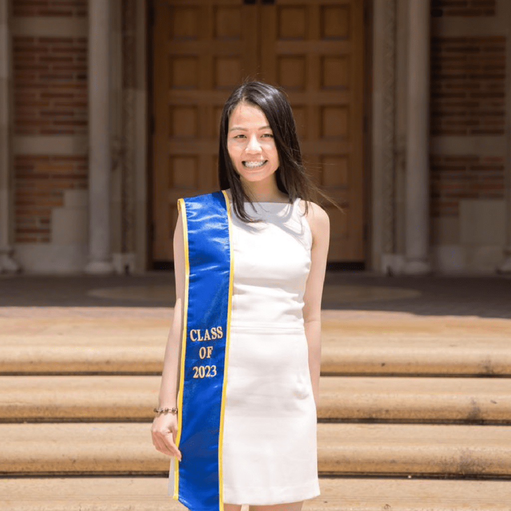 A young woman in a white dress stands on steps in front of large wooden doors, wearing a blue and gold sash that reads Class of 2023. She is smiling and posing for a graduation photo.