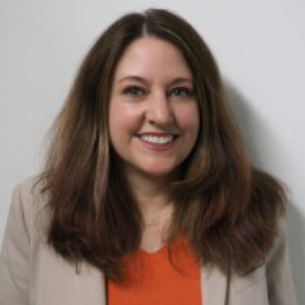 A woman with long, wavy brown hair, wearing a beige blazer over an orange top, smiles at the camera in front of a plain white background.