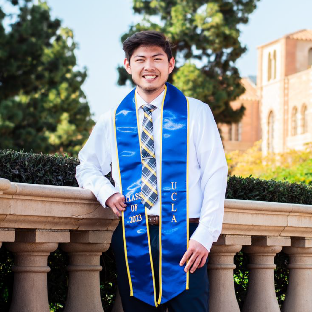 A young man wearing a blue UCLA graduation stole poses and smiles outdoors, leaning against a stone railing with trees and a campus building in the background.