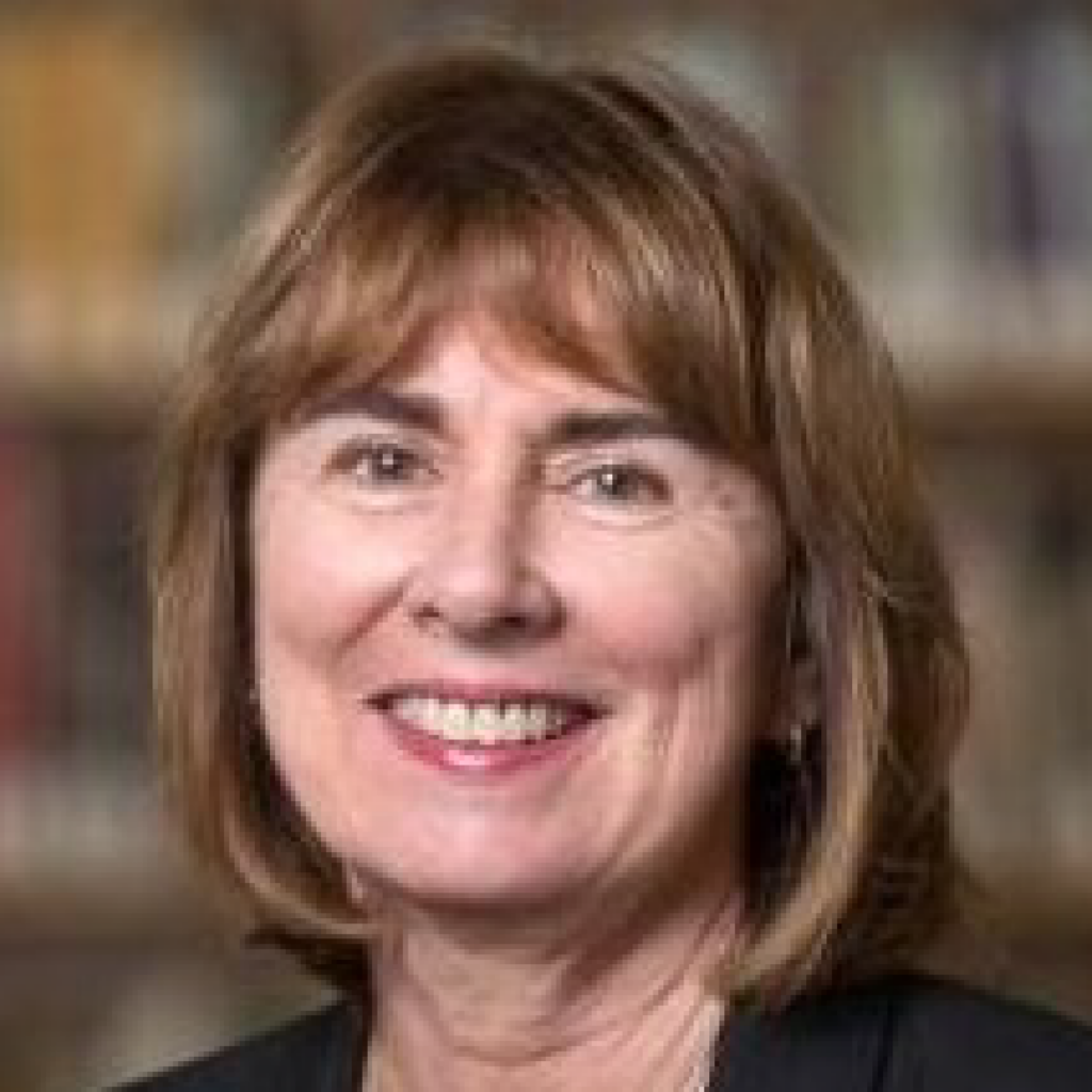 A middle-aged woman with light brown hair, smiling, wearing a dark jacket. The background is softly blurred, suggesting a bookshelf or library setting.