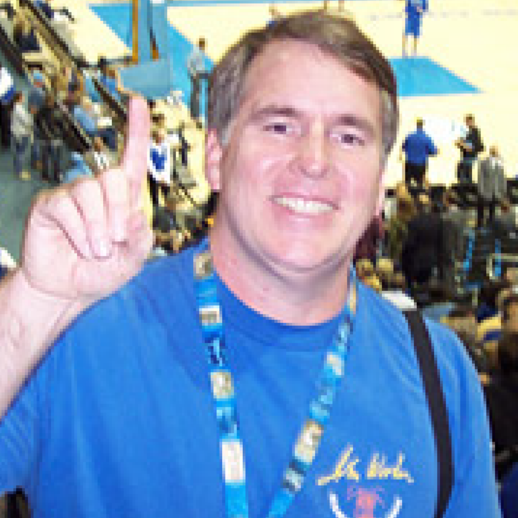 A smiling man wearing a blue shirt and event lanyard holds up one finger, standing in an indoor sports arena with people and a basketball court in the background.